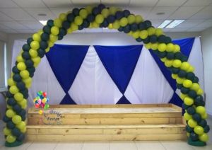 Arco de balões em espiral verde e amarelo tendo como fundo malha tensionada (fundo branco e triângulos azuis)